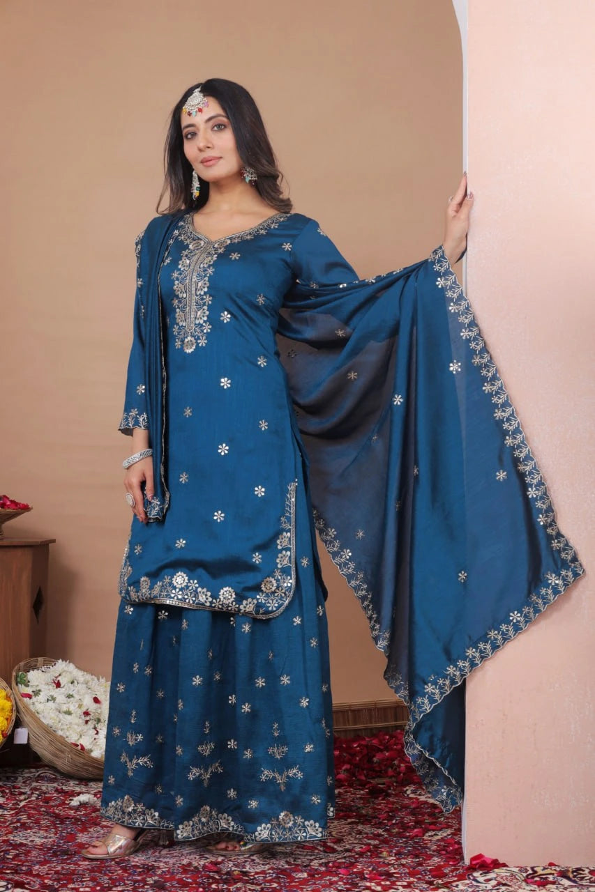 Woman wearing a blue embroidered traditional outfit with a dupatta against a beige wall.