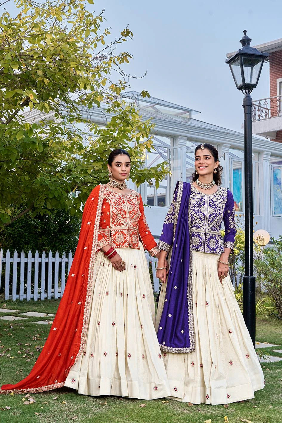 Two women in traditional outfits standing outdoors with greenery and a building in the background.