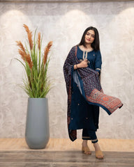 Woman in a blue traditional outfit standing next to a tall vase with plants against a light gray wall.