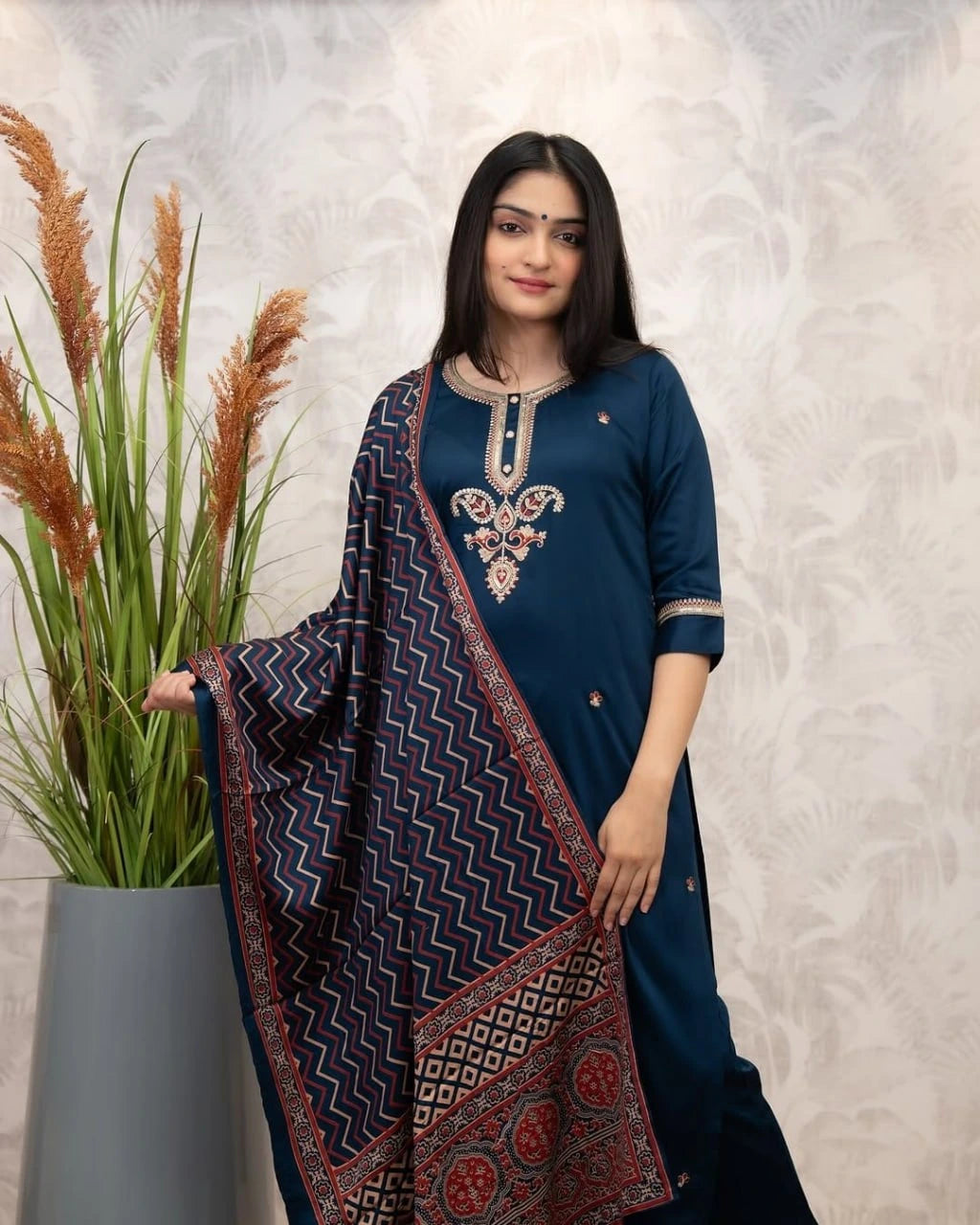 Woman in a blue traditional outfit with a patterned shawl, standing next to a plant against a light background