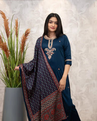 Woman in a blue traditional outfit with a patterned shawl, standing next to a plant against a light background