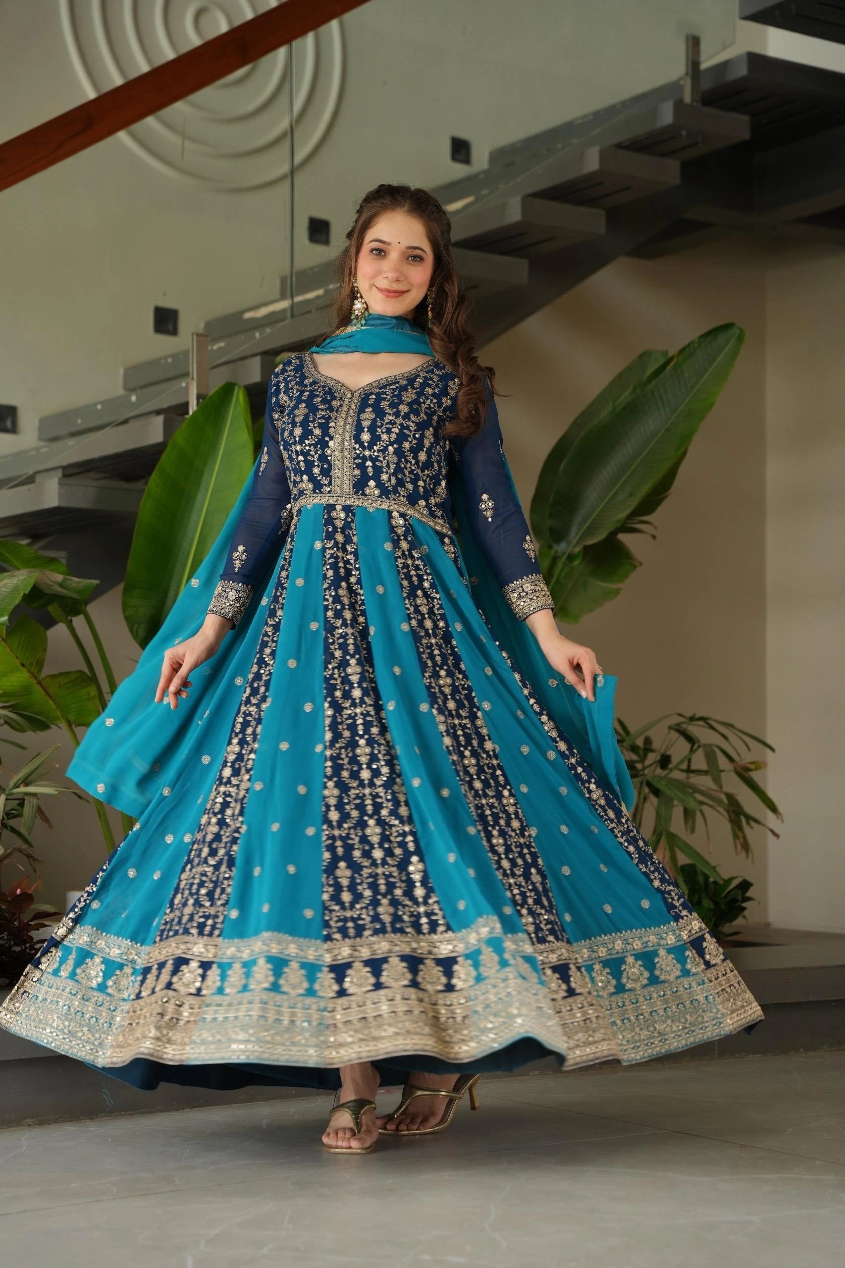 Woman in a blue and black embroidered traditional outfit standing indoors with plants in the background