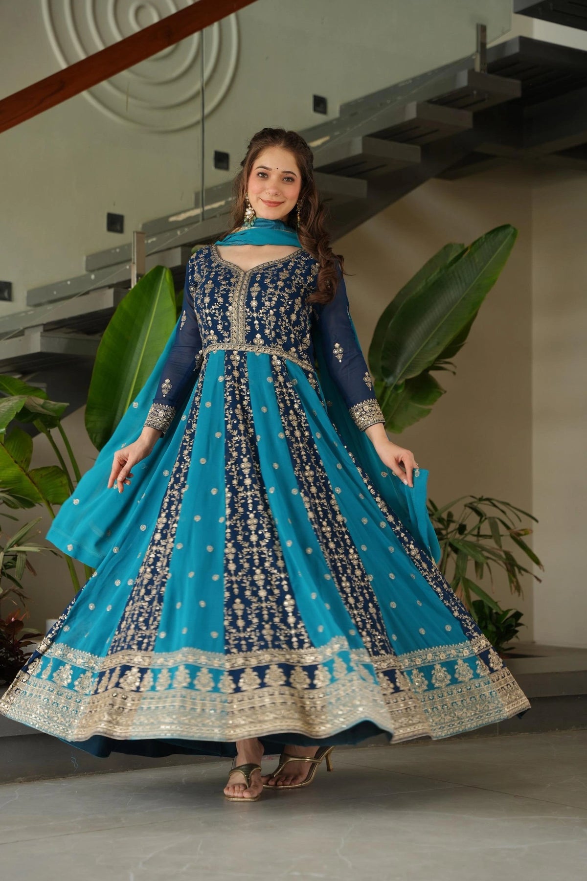 Woman in a blue and black embroidered traditional outfit standing indoors with plants in the background