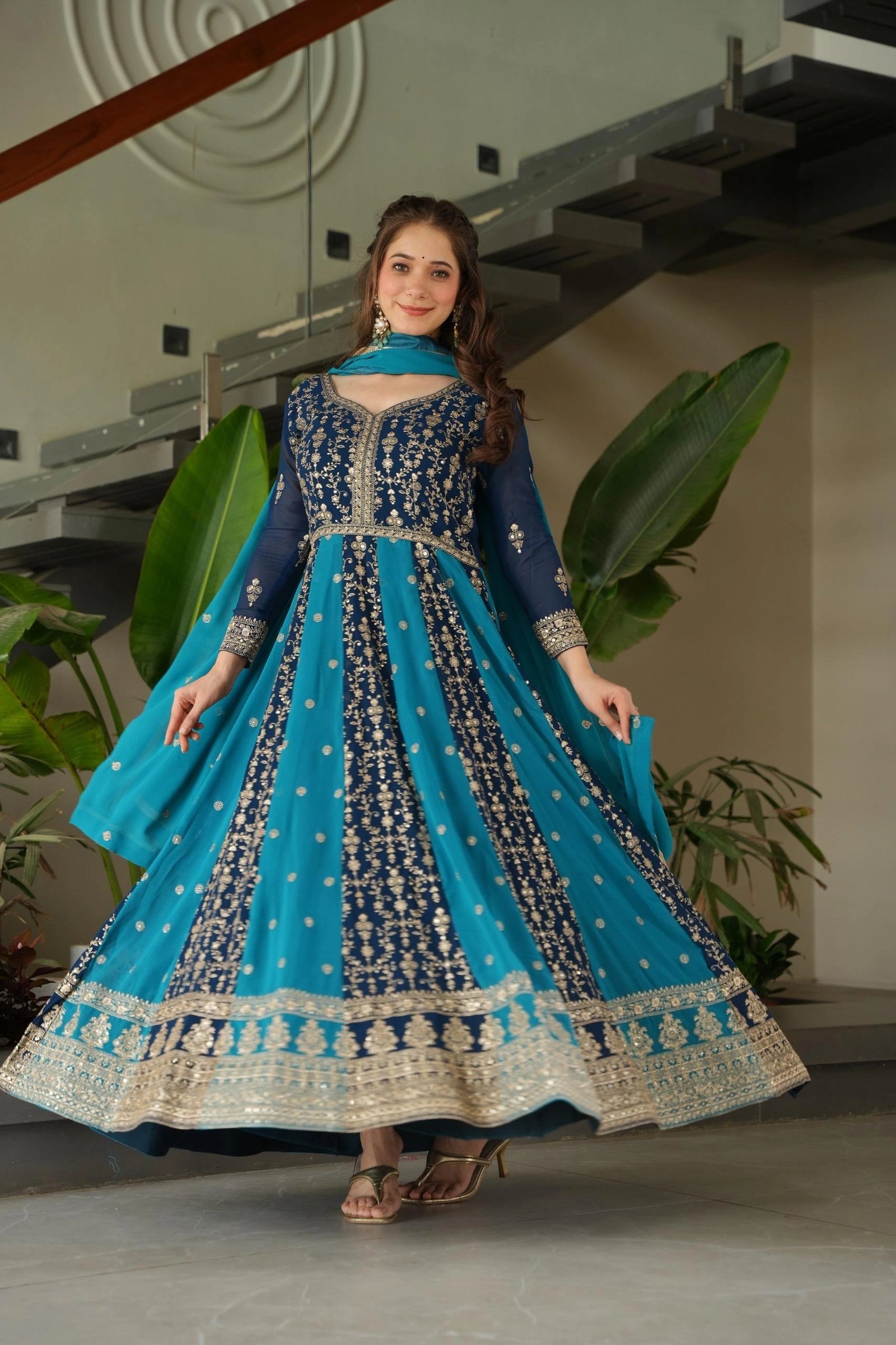 Woman in a blue and black embroidered traditional outfit standing indoors with plants in the background