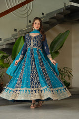 Woman in a blue and black embroidered traditional outfit standing indoors with plants in the background