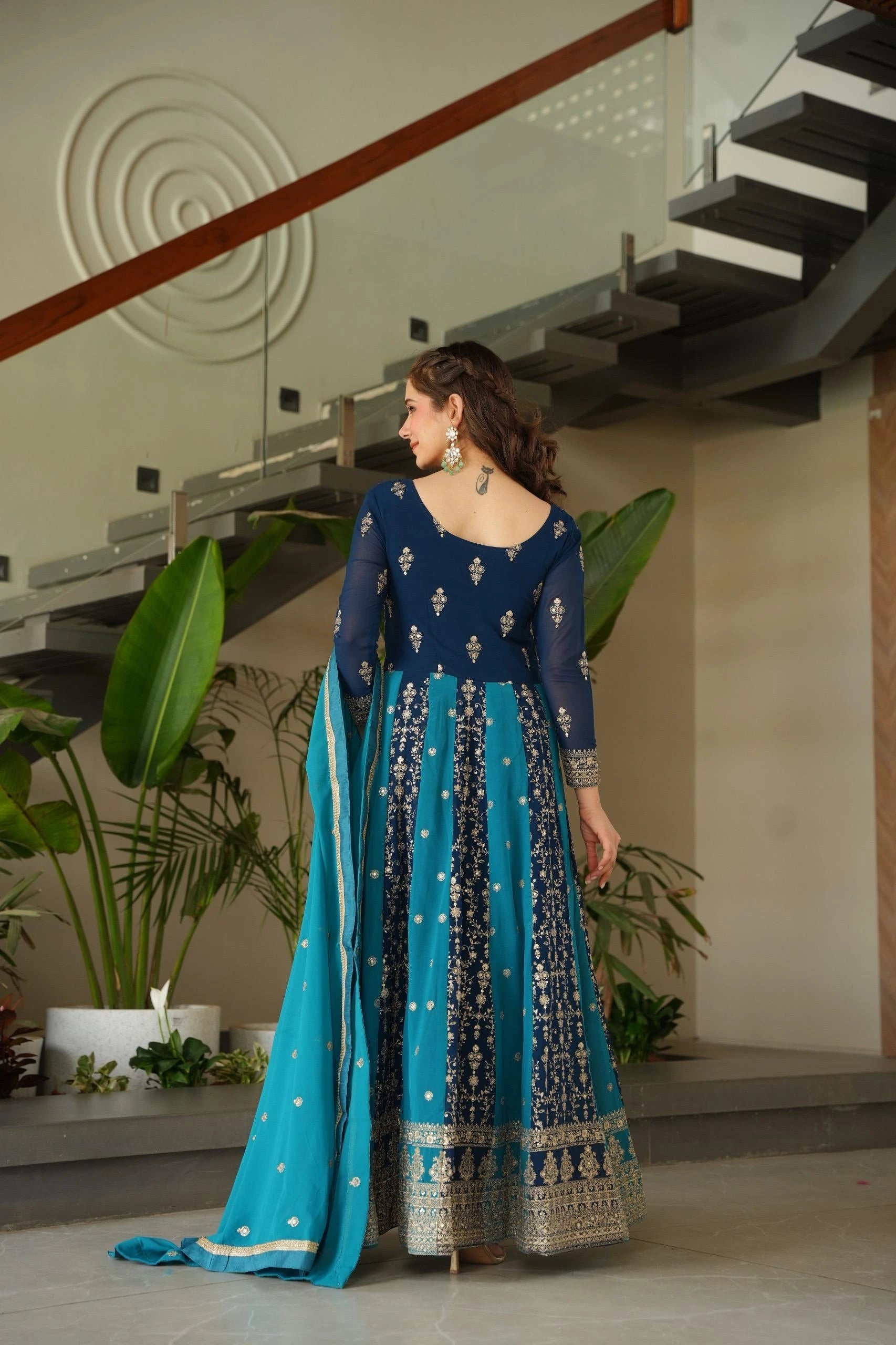 Woman in a blue and black embroidered traditional outfit standing indoors with plants and stairs in the background.