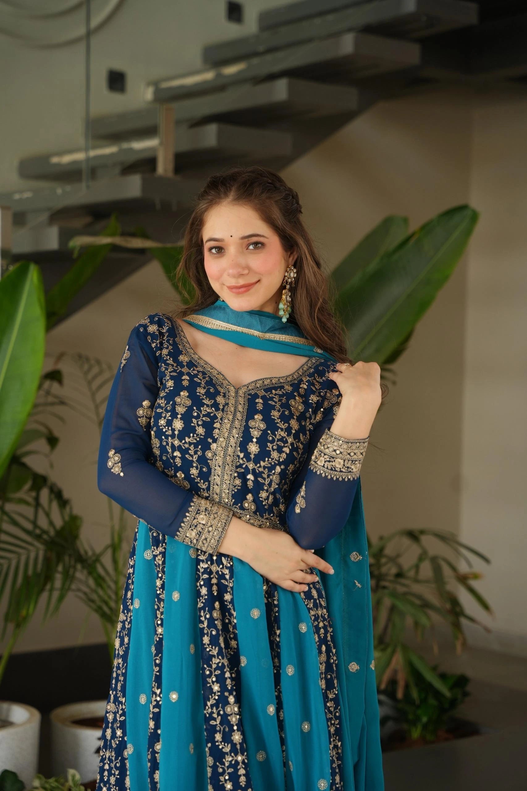 Woman in a blue and silver traditional outfit standing indoors with plants in the background