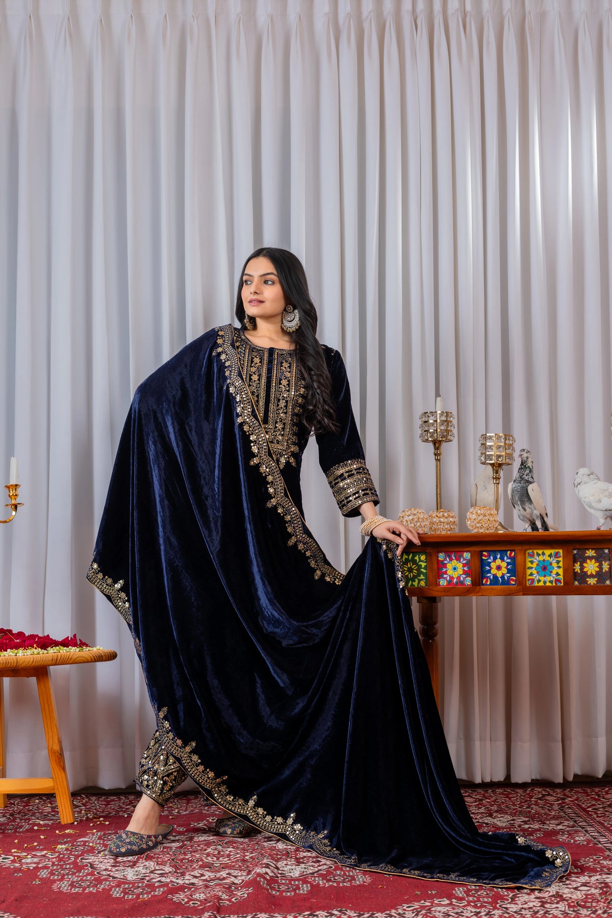 Woman in a navy blue embroidered outfit standing in a decorated room with a table and decorative items.