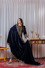 Woman in a navy blue embroidered outfit standing in a decorated room with a table and decorative items.
