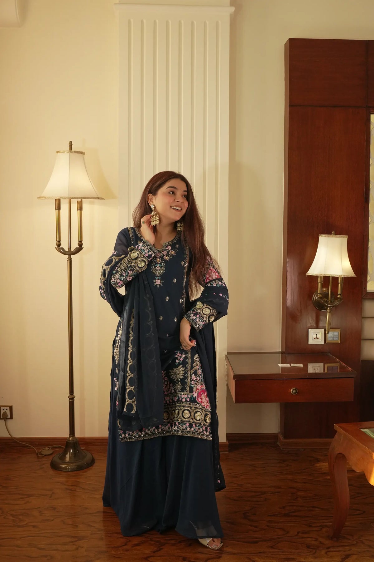 Woman in a dark blue embroidered dress standing in a room with a lamp and wooden furniture.