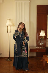 Woman in a dark blue embroidered dress standing in a room with a lamp and wooden furniture.