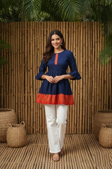 Woman wearing a blue and red top with white pants standing against a wooden panel background.