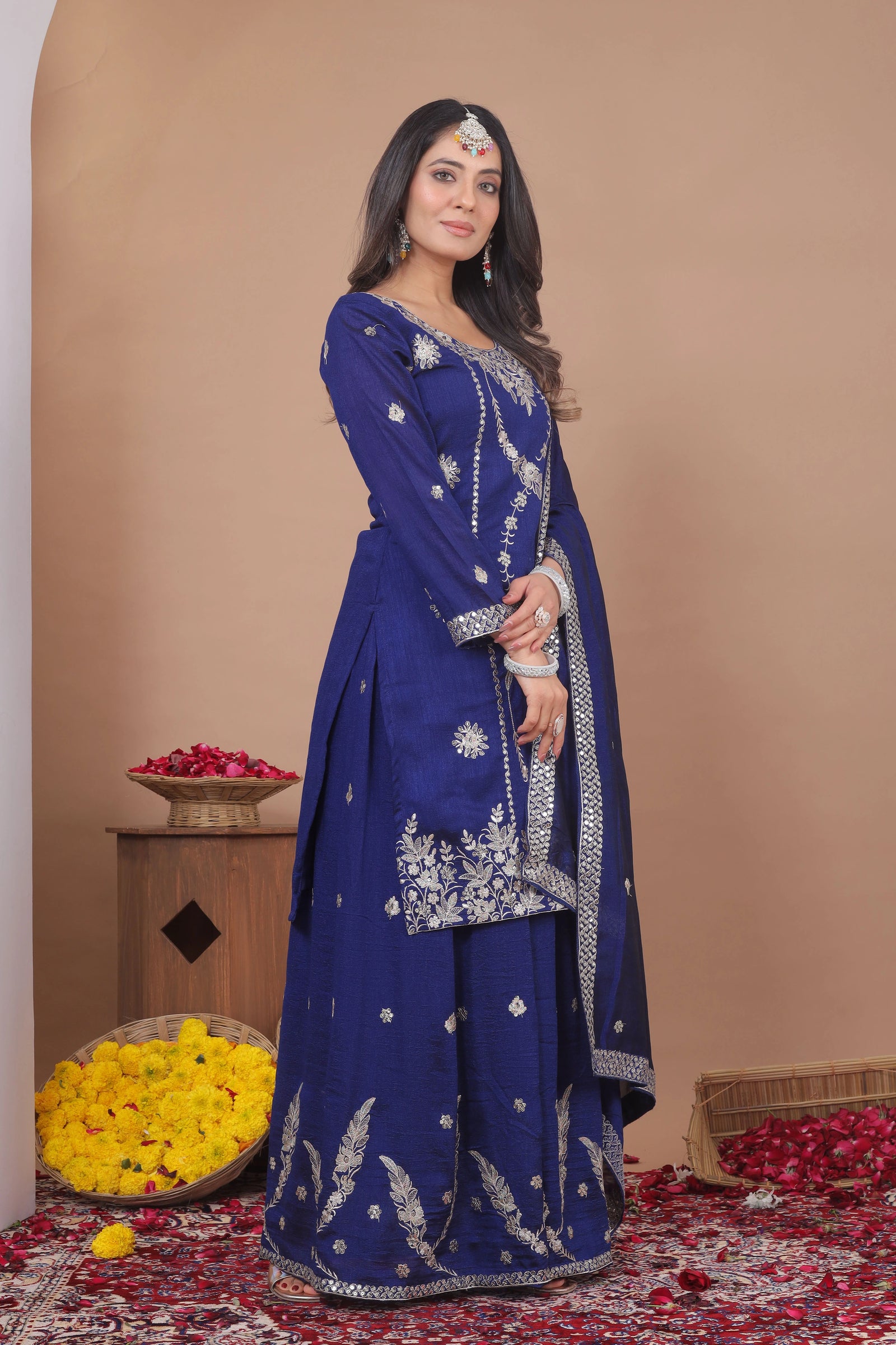 Woman in a blue embroidered traditional outfit standing against a beige wall with floral decorations on the floor.