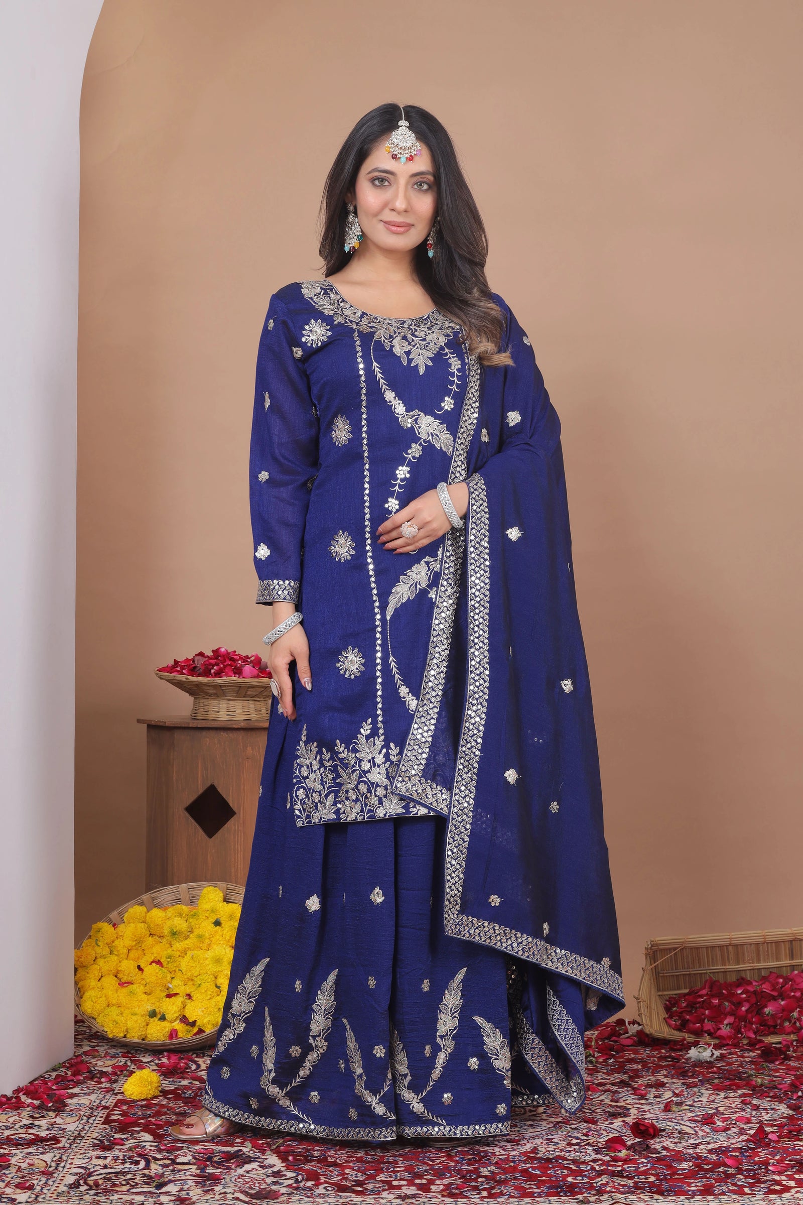 Woman in a blue embroidered traditional outfit standing against a beige wall with decorative elements.