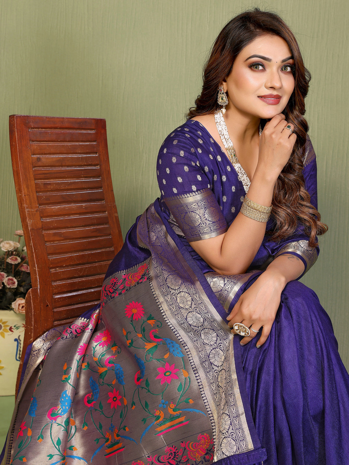 Woman wearing a purple saree with floral patterns, sitting against a green wall.