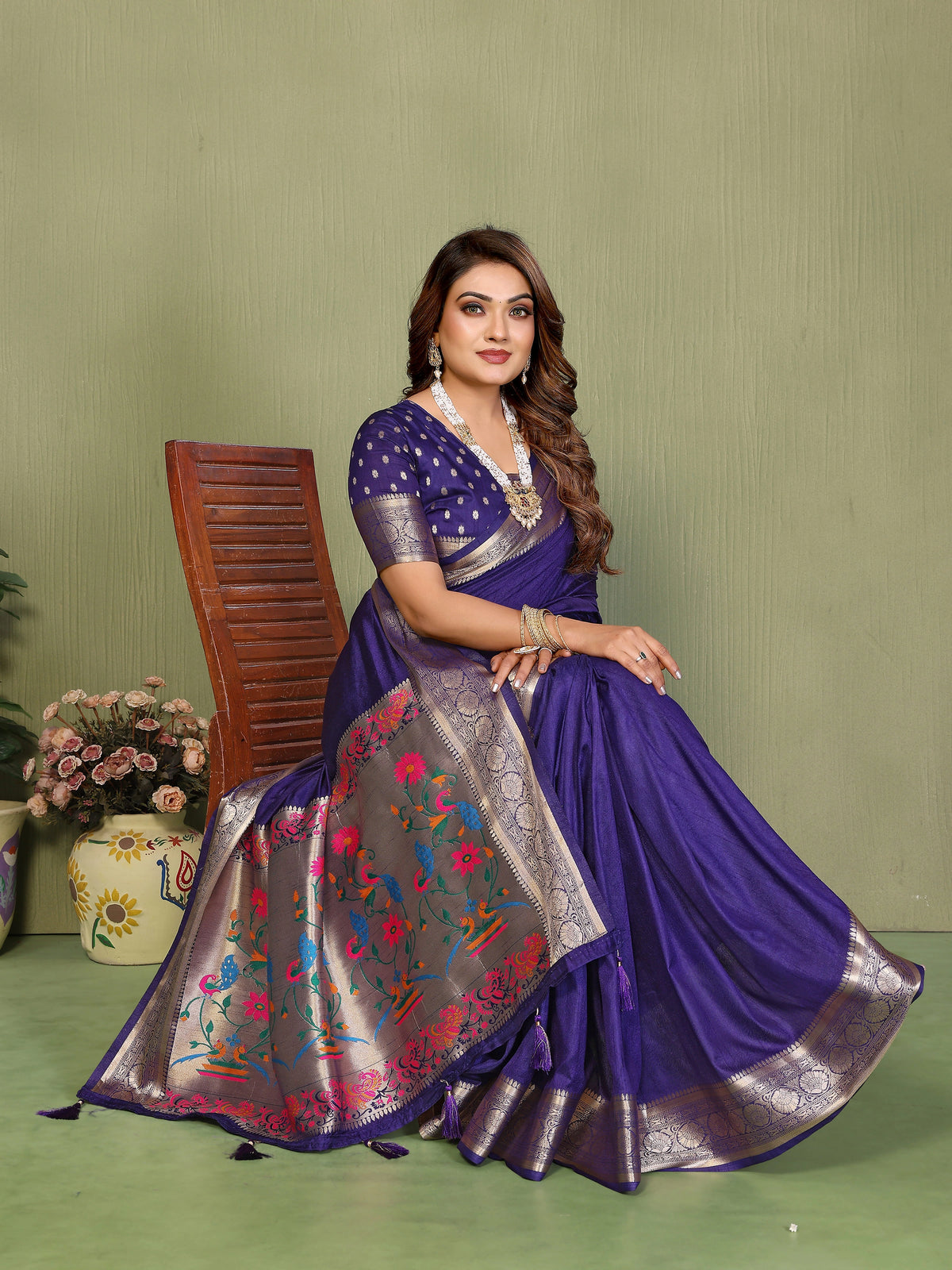 Woman in a traditional purple saree with floral patterns sitting on a chair against a green background