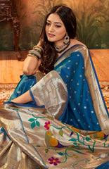 Woman wearing a blue and beige saree with floral and bird patterns, sitting on a wooden floor.