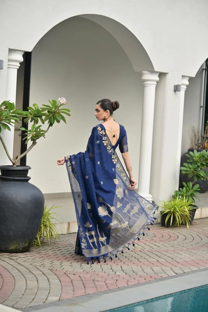 Woman in a blue saree standing in a courtyard with plants and arches.