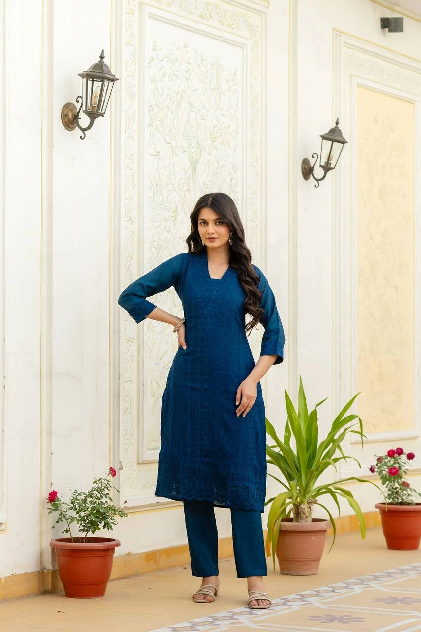 Woman in a blue outfit standing in a decorated room with plants and wall lights.