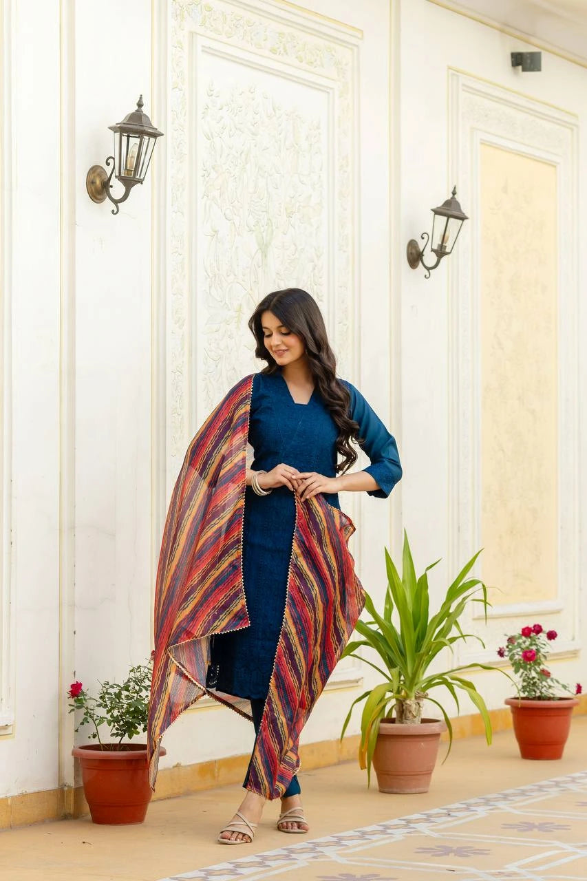 Woman in a blue outfit with a colorful scarf standing in a decorated room with plants.