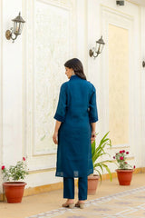 Woman in a blue kurta standing in a decorated room with plants and wall lights.