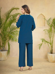 Woman in a blue outfit standing in a room with plants and a beige wall.