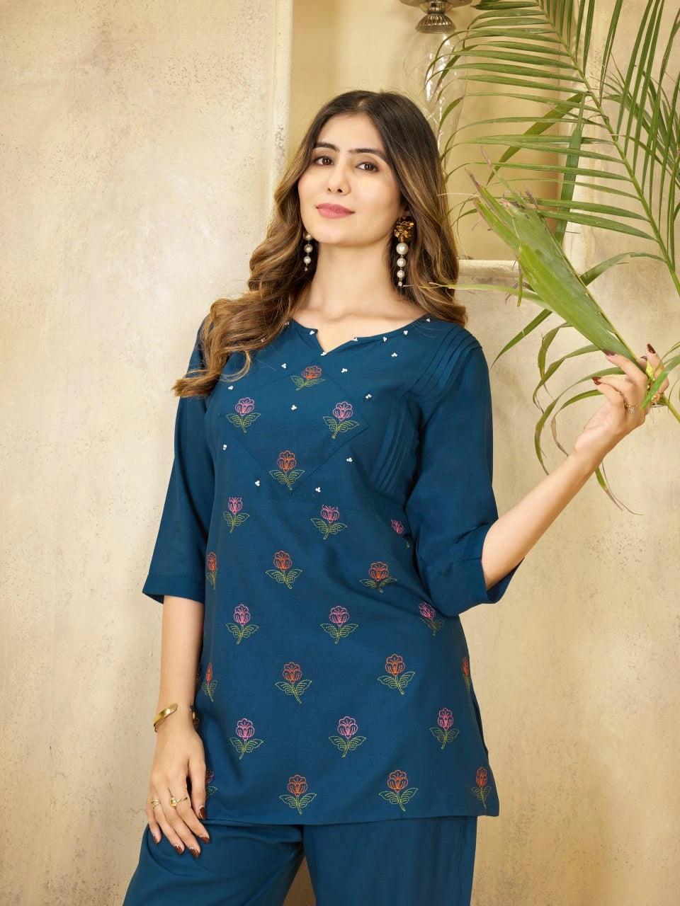 Woman wearing a blue embroidered top and pants holding a leaf against a beige wall.