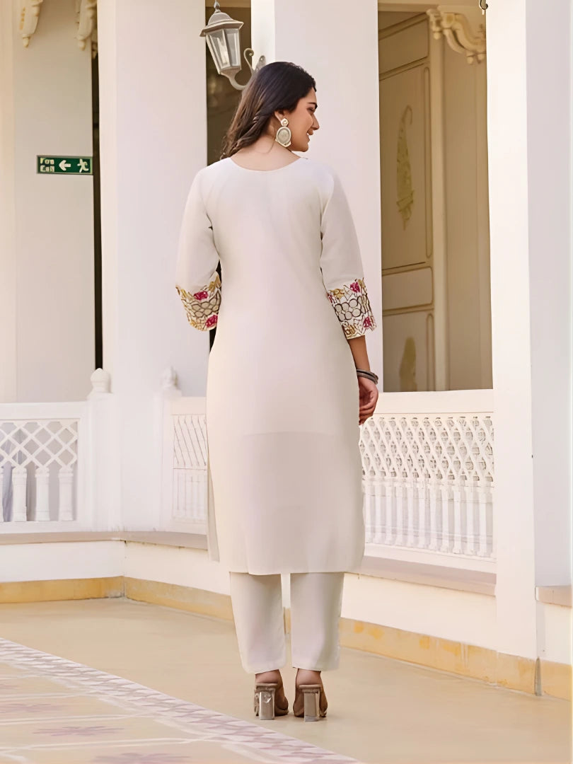 Woman in a white traditional outfit with embroidered sleeves standing in a decorative interior setting.