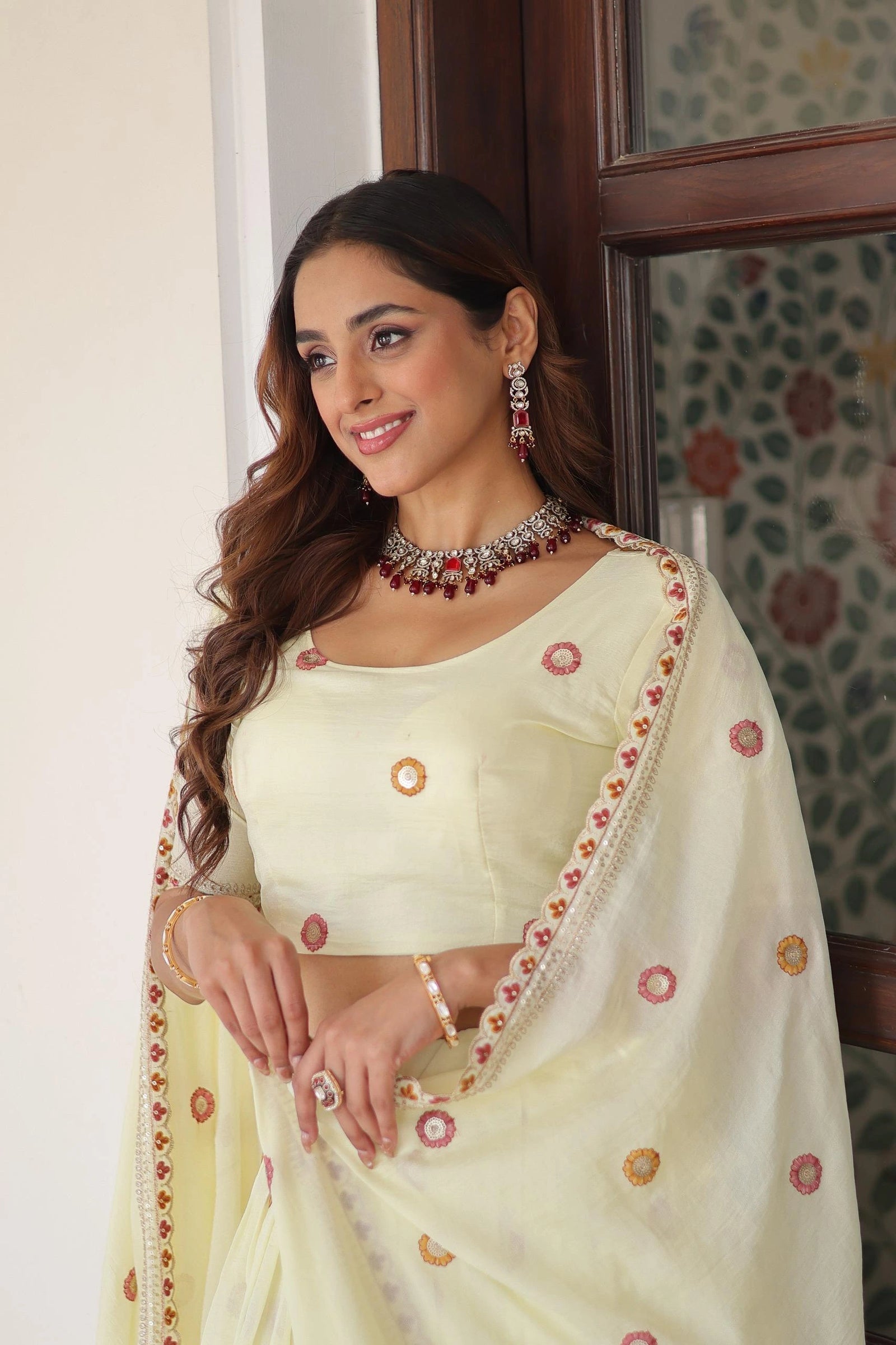 Woman wearing a cream saree with floral patterns in a room with patterned wallpaper.