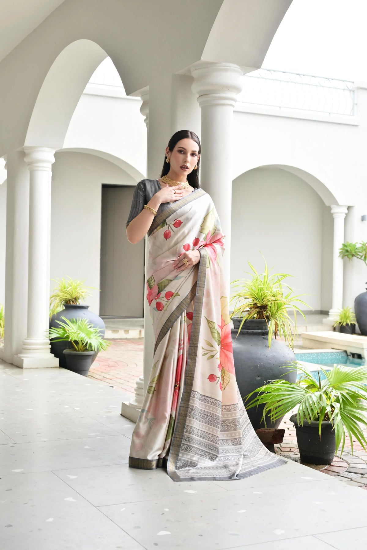 Woman in a floral saree standing in an outdoor setting with columns and plants.