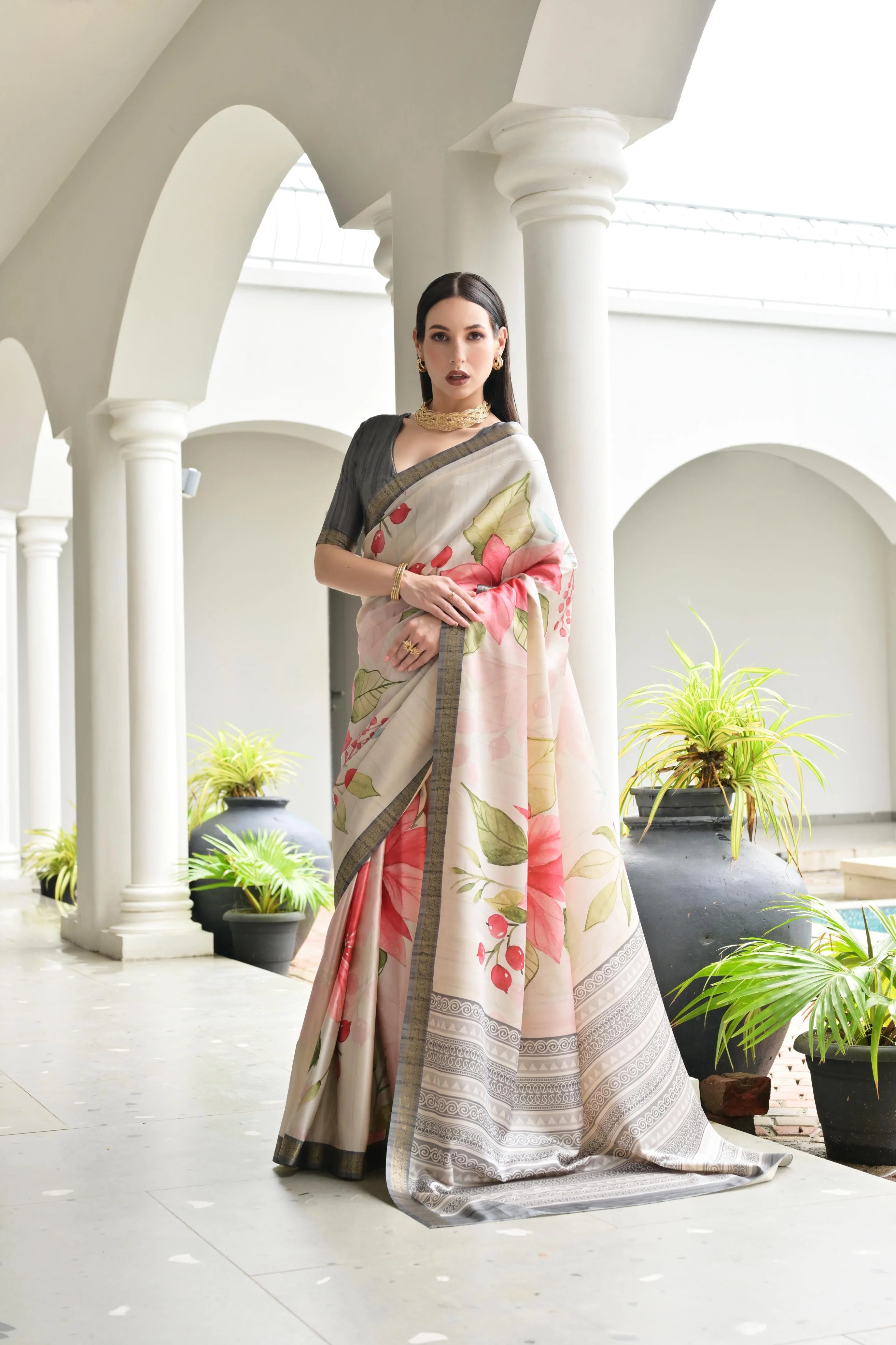 Woman in a floral saree standing in an elegant indoor setting with white columns and potted plants.