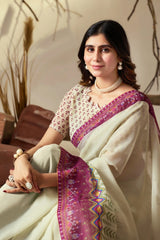 Woman in a white saree with purple border sitting indoors.