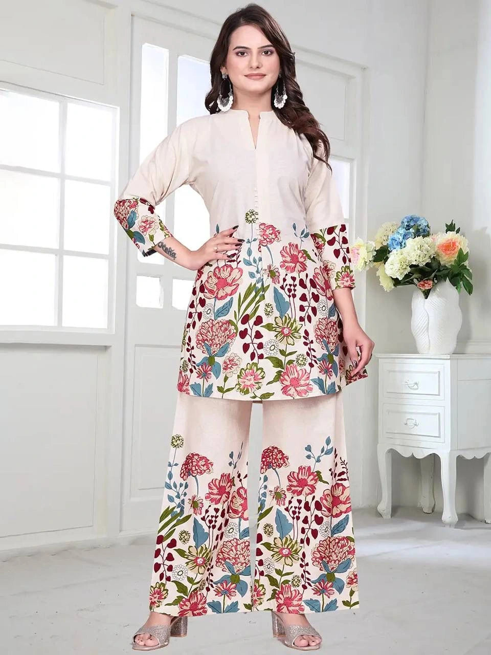 Woman wearing a floral outfit in a bright room with white walls and a vase of flowers.