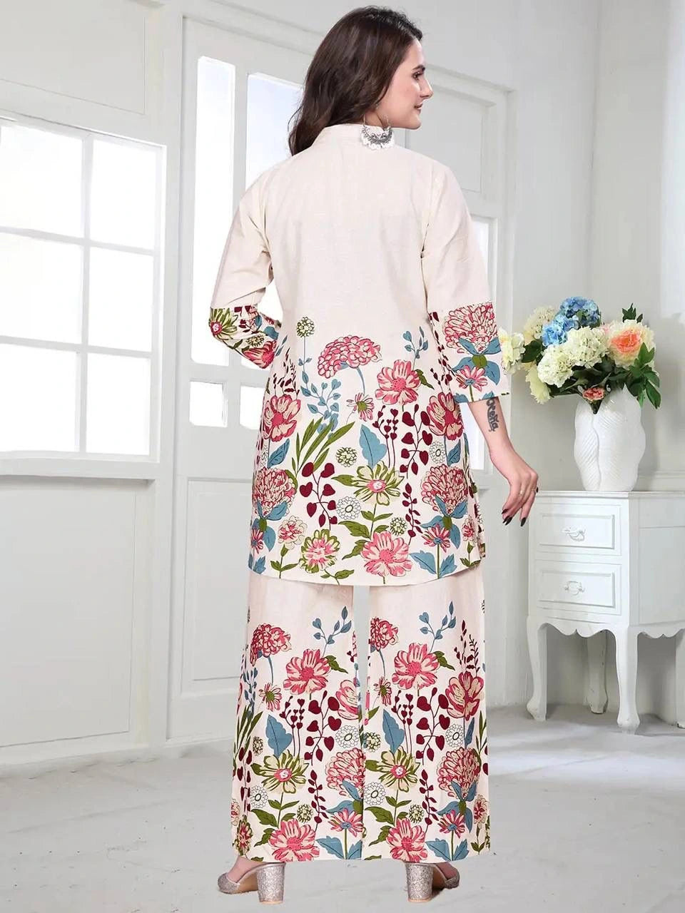 Woman wearing a floral dress in a bright room with white walls and a vase of flowers on a table.