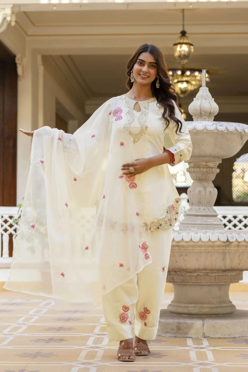 Woman in a white traditional outfit with floral patterns standing in an elegant outdoor setting.