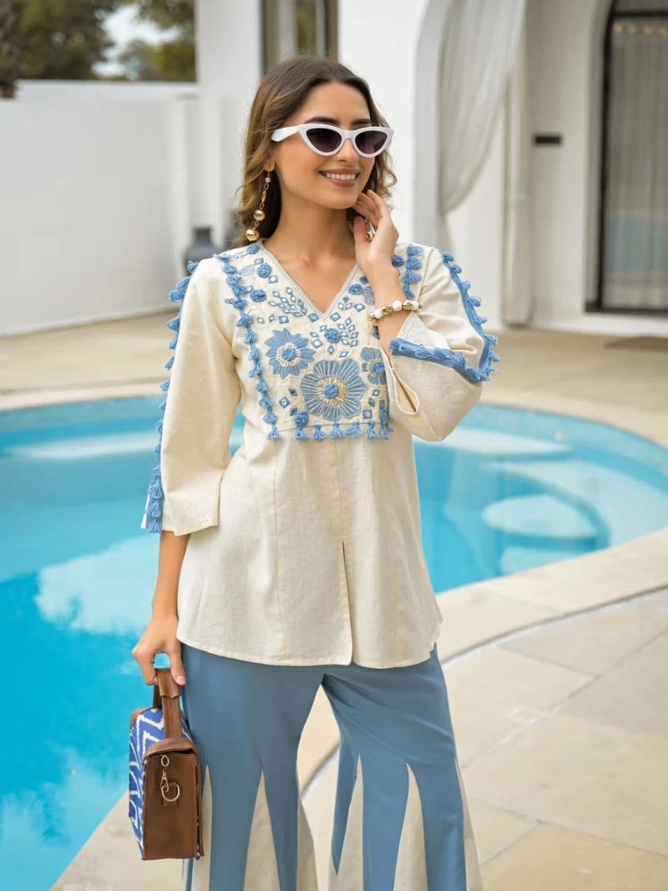 Woman in a white blouse with blue embroidery and blue pants standing by a pool.