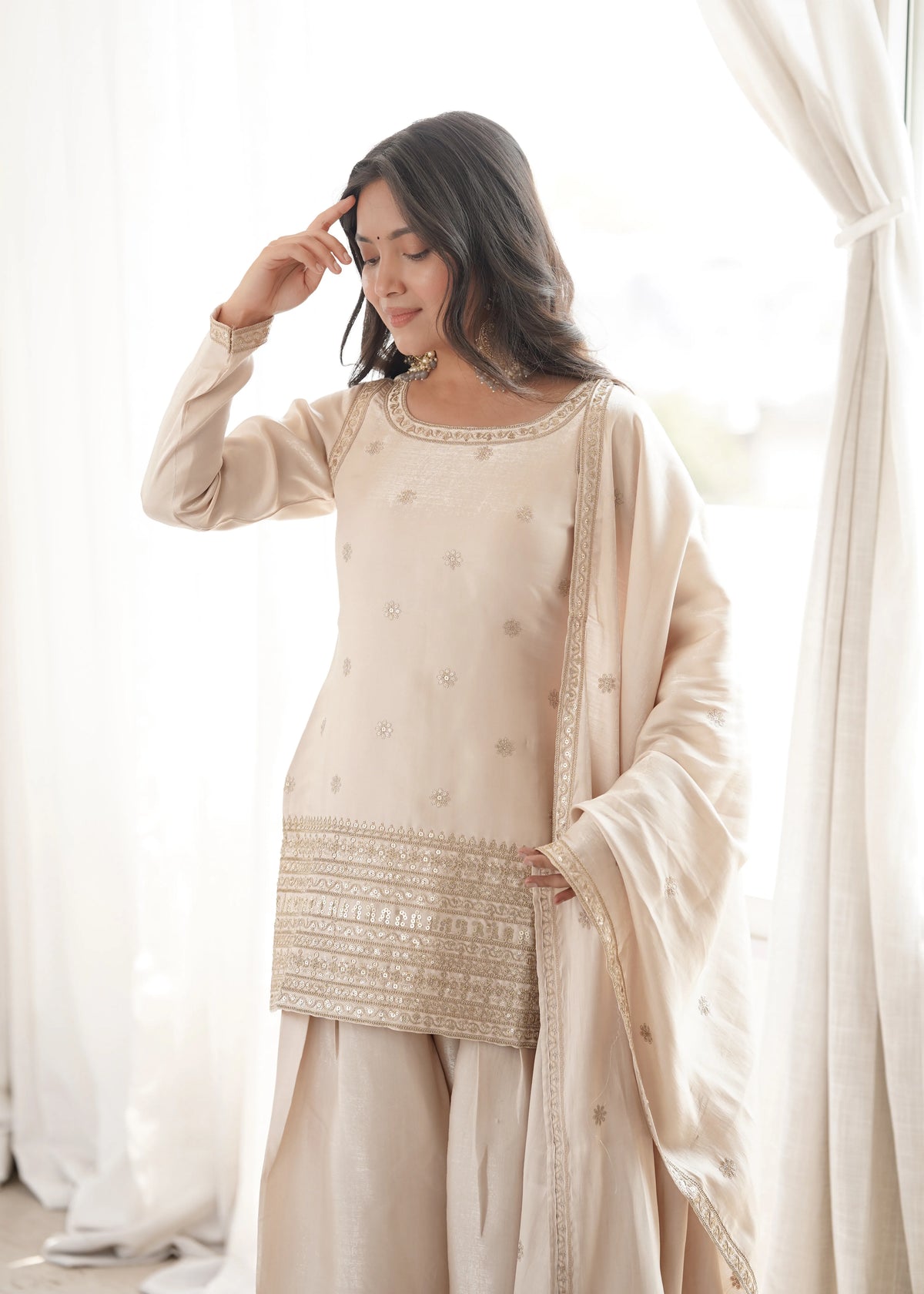 Woman wearing a beige traditional outfit with intricate patterns in a softly lit room.