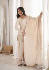 Woman in a beige traditional outfit standing in a softly lit room with curtains.