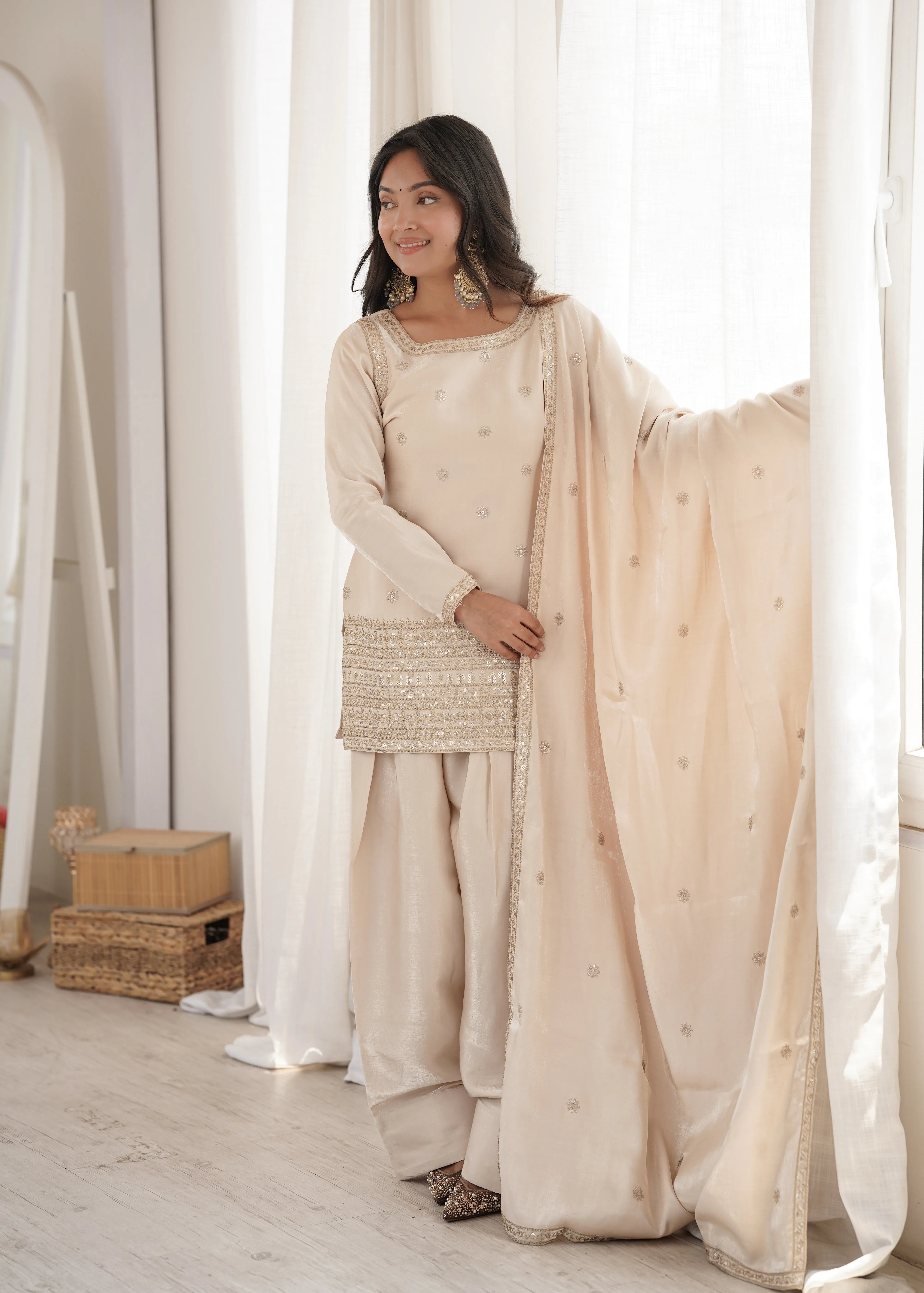 Woman in a beige traditional outfit standing in a bright room with white curtains.
