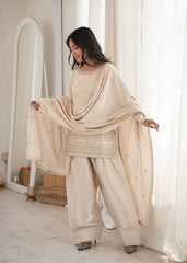 Woman in a beige traditional outfit standing in a room with white curtains.