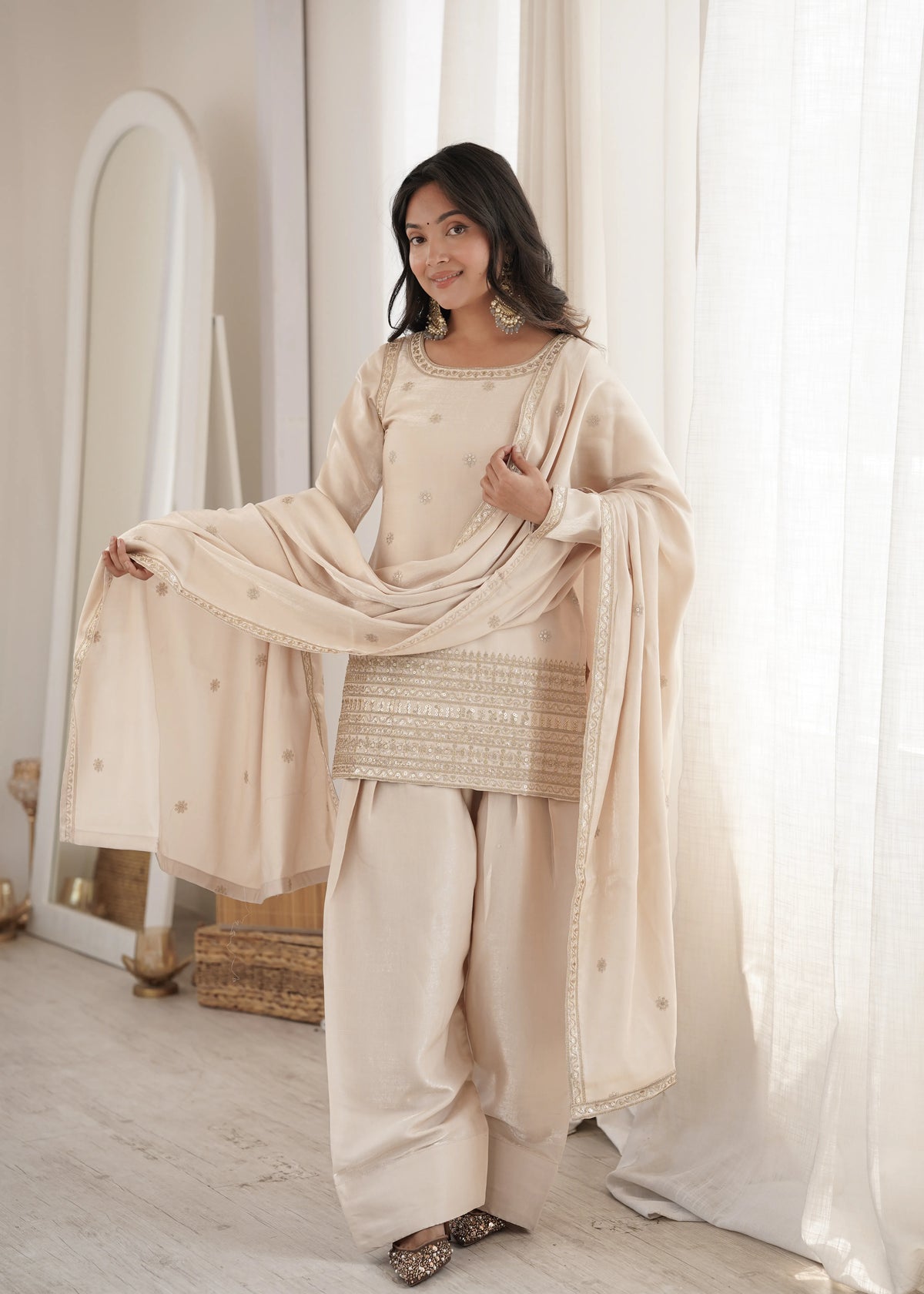 Woman in a beige traditional outfit standing in a room with white curtains.