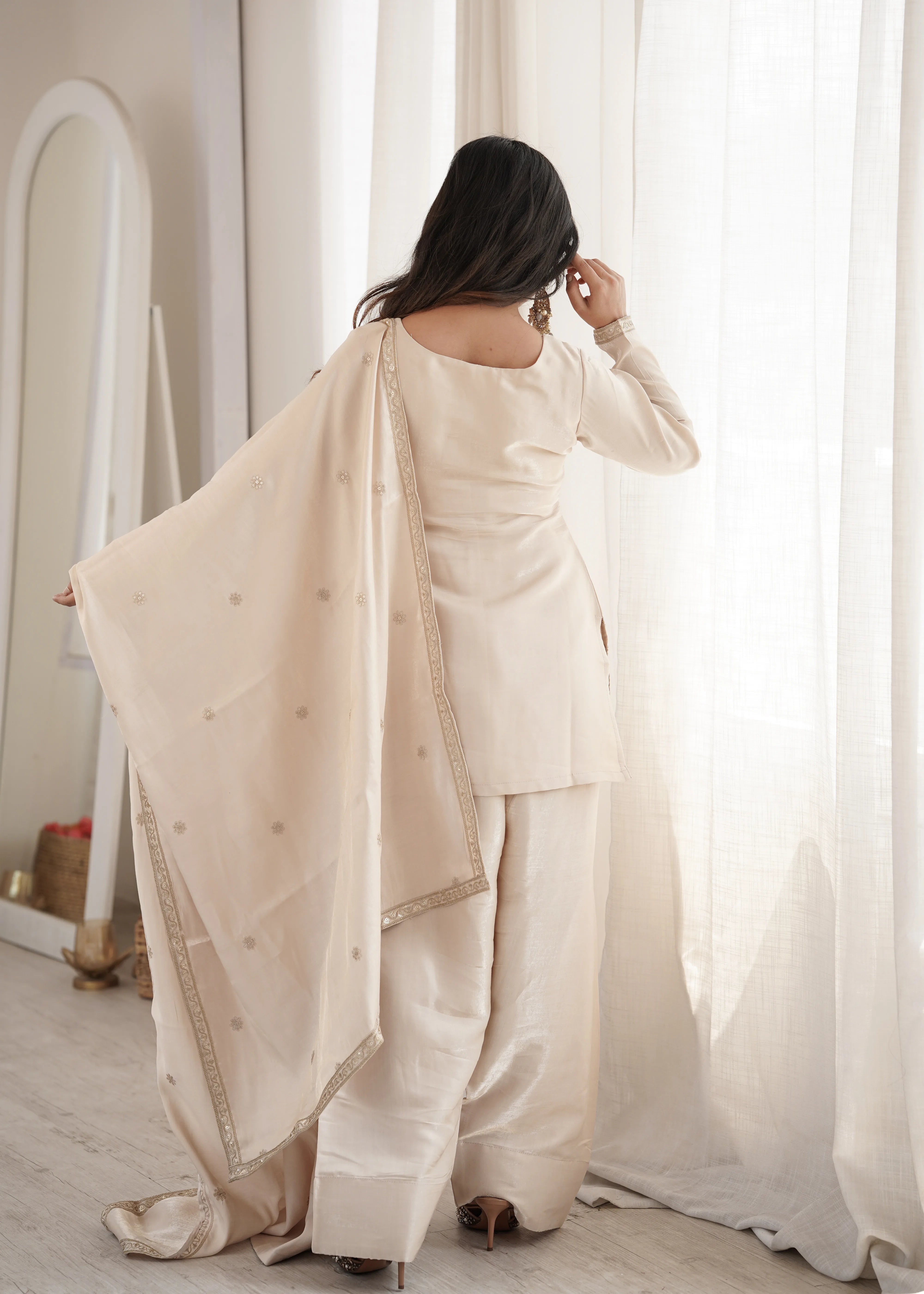 Woman in a beige traditional outfit standing in a softly lit room with white curtains.