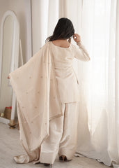 Woman in a beige traditional outfit standing in a softly lit room with white curtains.