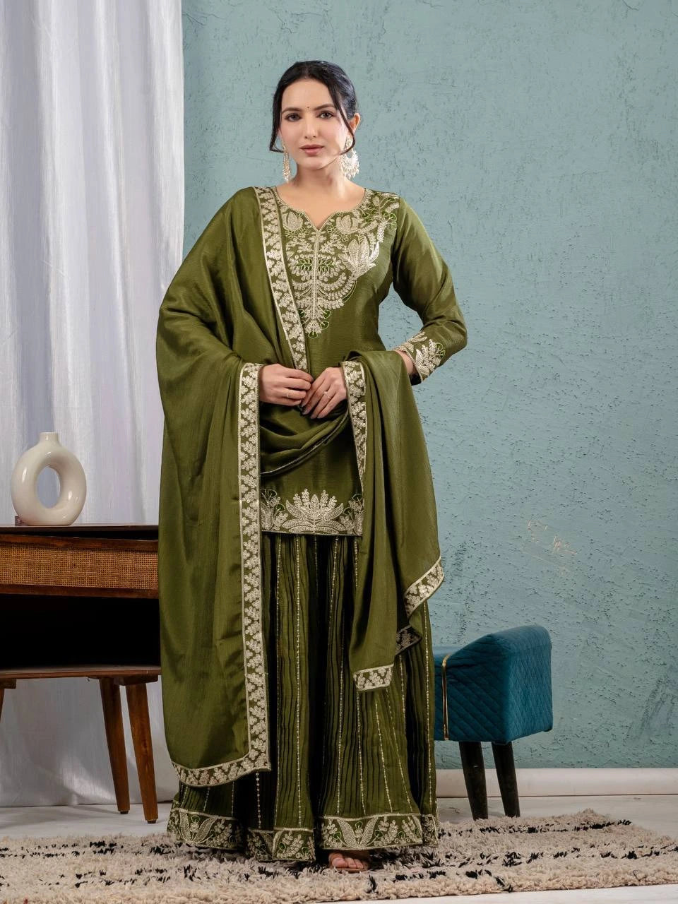Woman wearing a green traditional outfit with embroidery in a room with a blue wall and white curtains.