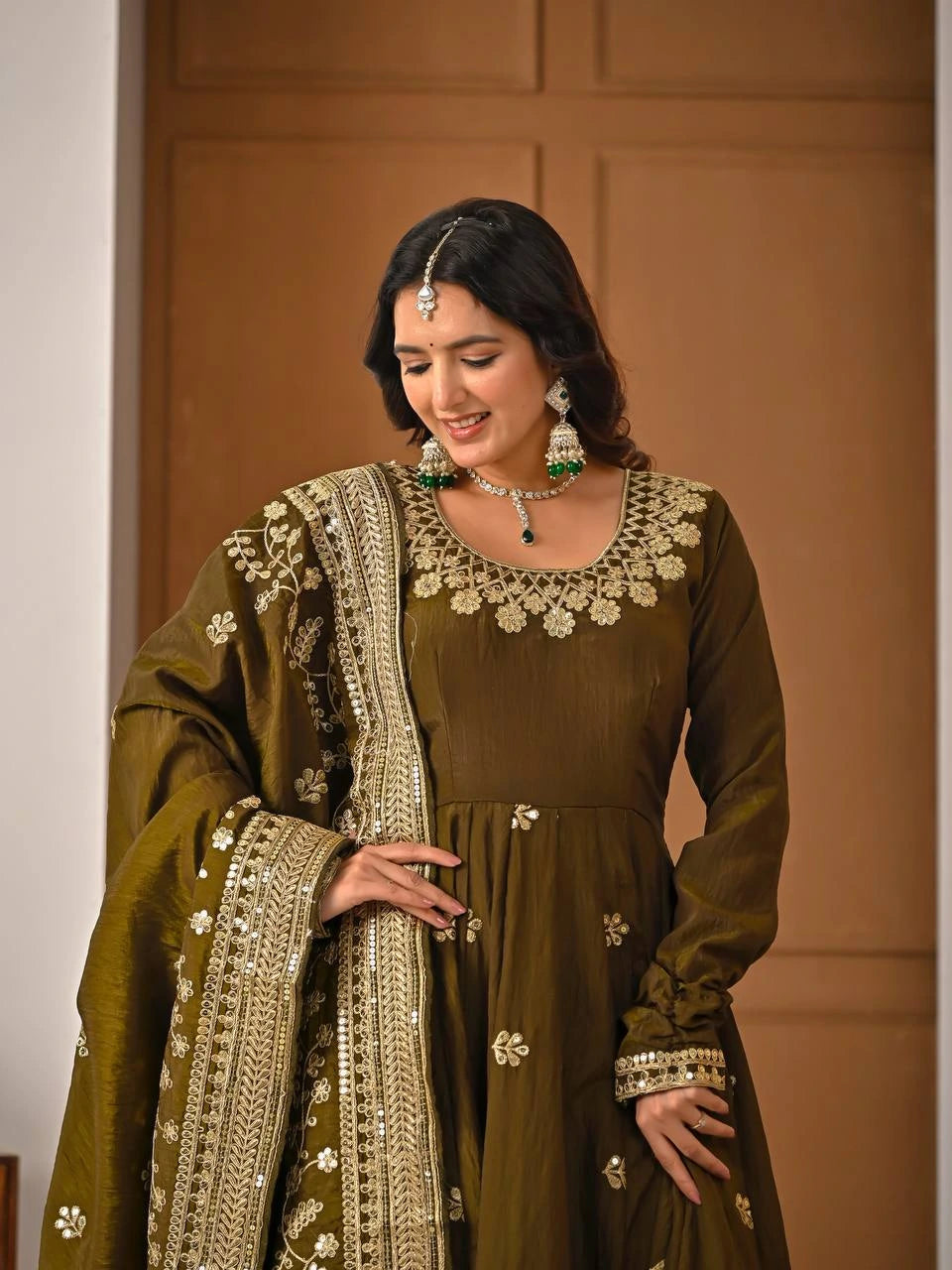 Woman in traditional olive green outfit with intricate patterns and jewelry against a brown background