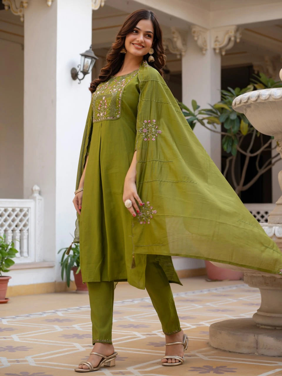 Woman in a green traditional outfit standing in an outdoor setting with plants and architectural elements.