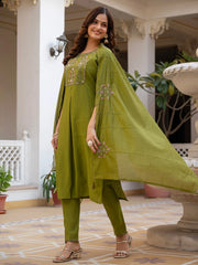 Woman in a green traditional outfit standing in an outdoor setting with plants and architectural elements.