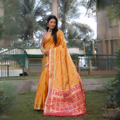 Woman wearing a yellow Banarasi silk saree adorned with gold zari motifs and a striking pink contrast pallu featuring traditional temple and floral patterns. She pairs the saree with a short sleeve pink blouse and accessorizes with gold jewelry, standing gracefully on a manicured lawn in an outdoor garden setting with fencing, lush greenery, and tropical trees in the background.