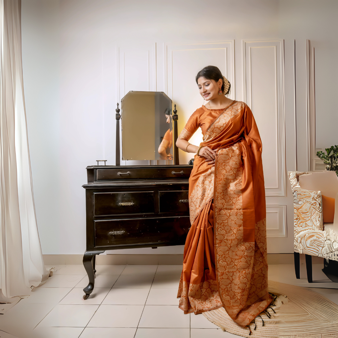 A woman models a luxurious Terracotta Orange Raw Silk Saree. The saree features a wide border and pallu with dense, tone-on-tone gold Zari weaving in a Kalamkari-style floral and paisley pattern. She wears a matching short-sleeve orange blouse and subtle gold jewelry, posing indoors next to a dark wooden vanity.