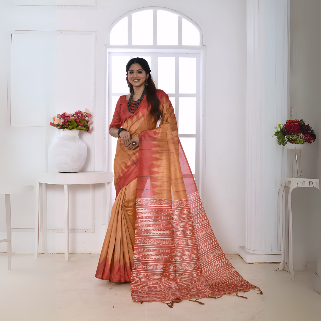 A woman models a graceful Copper Orange Soft Tussar Silk Saree. The saree features a zigzag pattern where the body meets the pallu, and the pallu has a wide panel of dense white and red geometric tribal-style print. She wears a coral blouse and a chunky dark beaded necklace, posing indoors by a large window.
